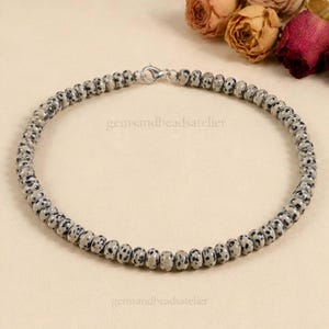 May include: A necklace composed of small, round, beige beads with black spots, resembling dalmatian jasper. The beads are strung in a circular pattern and secured with a silver-toned metal lobster claw clasp. The text gemsandbeadsatelier is visible.