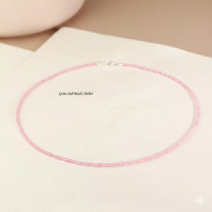 Puede incluir: Un delicado collar de cuentas rosas con un cierre plateado. El collar está compuesto por pequeñas cuentas redondas y se muestra sobre una superficie blanca. El texto "Gems and Beads Atelier" es visible.