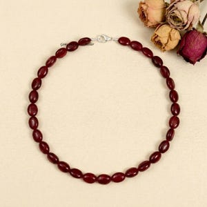 May include: A necklace composed of oval-shaped, dark red beads. The beads are strung together, forming a complete circle. A silver-colored clasp secures the necklace. The background is a light beige color, with dried flowers in the upper right corner.