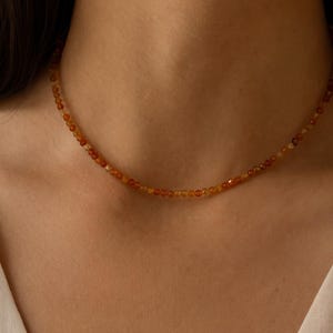 Puede incluir: Un delicado collar compuesto por pequeñas cuentas facetadas en tonos naranja y amarillo. El collar se lleva alrededor del cuello, mostrando los tonos cálidos de las piedras preciosas. La joya es un accesorio sencillo y elegante.