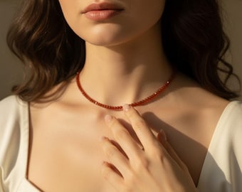 May include: A delicate red beaded necklace. The necklace is worn around the neck. The model is wearing a white top. The necklace is made of small, round beads. The beads are a deep red color.