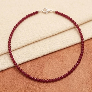 May include: A fine necklace composed of small, faceted, deep red beads. The beads are strung together, forming a complete circle, and are secured with a silver-coloured clasp. The necklace is displayed on a neutral-toned fabric.