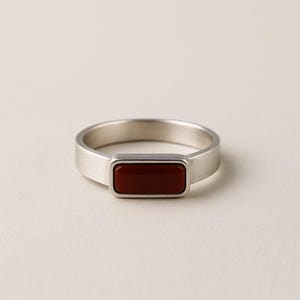 Könnte beinhalten: Ein silberner Ring mit einem rechteckigen, dunkelroten Stein. Der Ring hat eine gebürstete Metalloberfläche und ein schlichtes, modernes Design. Der Stein ist in einer Lünettenfassung gefasst, und das Band ist glatt und breit.