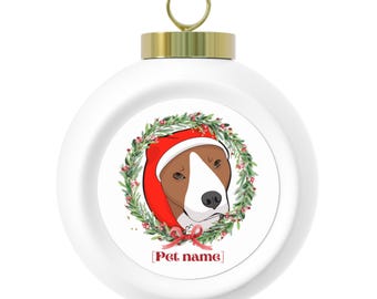 May include: White Christmas ornament featuring a dog wearing a Santa hat and framed by a green wreath with red berries. The ornament has a gold cap and a loop for hanging. The text '[Pet name]' is below the dog's image.