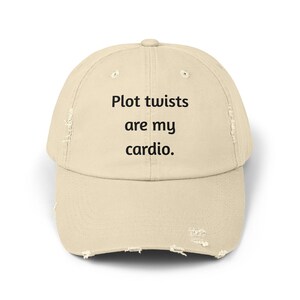 Puede incluir: Gorra de béisbol beige desgastada con el texto "Plot twists are my cardio" en negro. La gorra tiene una visera curva y un aspecto desgastado, lo que sugiere un estilo casual.