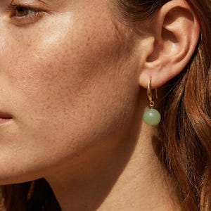 May include: A close-up shot of a person's face, with a focus on a gold hoop earring with a light green, round stone. The person has fair skin, freckles, and reddish-brown hair. The background is a solid, neutral color.