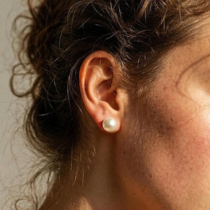 May include: A single round, smooth, off-white pearl stud earring worn in the earlobe. The pearl has a lustrous, reflective surface and a spherical shape, positioned centrally against the skin of the ear, showcasing a classic jewelry design.
