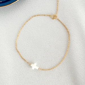 May include: A delicate gold-coloured chain bracelet featuring a small, white cross pendant. The bracelet is displayed on a light-coloured surface, showcasing its minimalist design and elegant simplicity. The cross is centered on the chain.