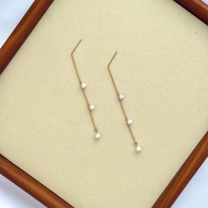 May include: A pair of gold-coloured earrings. Each earring has a fine chain with three small, white pearls. The earrings are presented on a beige surface within a wooden frame, highlighting their elegant design.