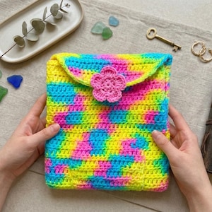 May include: A rectangular crochet pouch featuring a vibrant tie-dye pattern in neon yellow, bright pink, and electric blue. The pouch has a fold-over flap secured by a single, solid pink crocheted flower button centered on the front exterior.