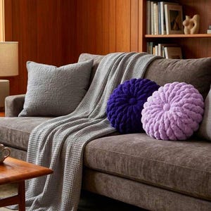 May include: A brown sofa featuring a grey quilted throw blanket, a large grey square pillow, and two round textured pillows in deep purple and light lavender. The sofa is positioned in front of a wooden wall with a bookshelf containing various books.
