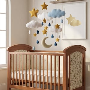 May include: A wooden crib with floral side panels and a white mattress, positioned beneath a hanging mobile featuring felt stars, clouds, a crescent moon, and blue and white raindrops suspended from a circular wooden frame by thin white strings.