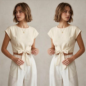 May include: Cream-colored linen crop top with a bow detail at the waist. The top has short sleeves and a round neckline. Paired with high-waisted, wide-leg white linen pants. A gold necklace is visible.