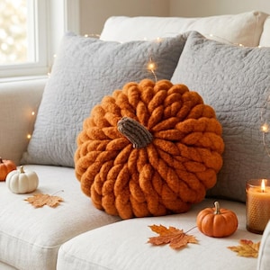 May include: A round, orange, chunky knit pumpkin-shaped cushion with a brown textured stem. The cushion is displayed alongside a small white pumpkin, a small orange pumpkin, a glass candle holder with a lit candle, and scattered dried autumn leaves.
