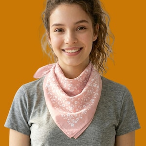 May include: A pink bandana with a white floral pattern is tied around the neck. The square scarf is made of soft fabric and is folded into a triangle. The background is a solid orange color.