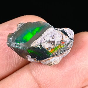 May include: A raw black opal gemstone, displaying vivid green and orange hues. The opal is held in a hand, highlighting its natural, unpolished state. The stone has a dark base colour with areas of iridescent play-of-colour.