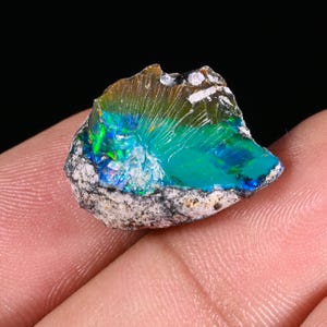 Op de afbeelding: Een levendige, ruwe opaal edelsteen met een opvallende kleurenweergave. De opaal heeft een mix van tinten van blauwgroen, blauw en groen, met flitsen van iriserende kleuren. De steen is geplaatst tegen een donkere achtergrond, wat de natuurlijke schoonheid benadrukt. De opaal wordt in een hand gehouden.