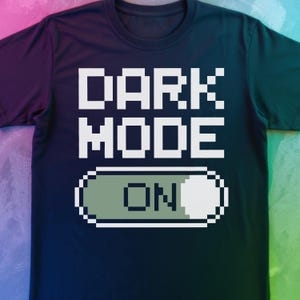 May include: Dark blue t-shirt with the words "DARK MODE" in white, pixelated font. Below the text is a pixelated switch with the word "ON" in white. The shirt has a crew neck.