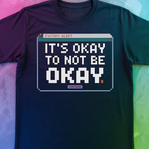 May include: Dark blue t-shirt featuring a pixelated design with the text "IT'S OKAY TO NOT BE OKAY." The graphic includes "SYSTEM ALERT" and "CONTINUE" in a retro computer style, set against a dark blue background.