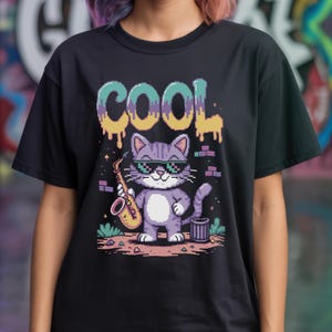 Peut inclure: T-shirt noir avec une illustration pixélisée d'un chat cool portant des lunettes de soleil et tenant un saxophone. Le mot "COOL" est imprimé au-dessus du chat en lettres colorées dégoulinantes. Le chat est à côté d'une poubelle.