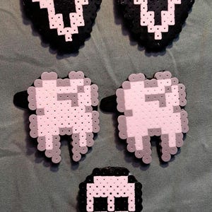 May include: Five pixel art accessories made from fused plastic beads. Two pieces are white and black ghost masks, two are white and grey teeth, and one is a white and black skull with crossbones. Each piece features a black clip on the back.