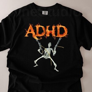 Könnte beinhalten: Schwarzes T-Shirt mit dem Wort "ADHS" in orangefarbenen Flammen über einer Skelettfigur, die Waffen hält. Das Design ist auf dem T-Shirt zentriert, mit dem Text und der Grafik in einem auffälligen Stil.