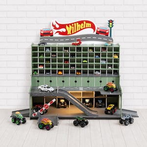 Può includere: Un garage per auto giocattolo a più livelli, verde e grigio, con una rampa, parcheggi e un cartello con la scritta "Wilhelm". Il garage è pieno di macchinine giocattolo, tra cui monster truck e auto da corsa. È visibile anche un semaforo.