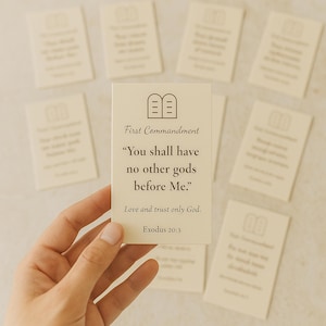 Puede incluir: Una mano sostiene una tarjeta de color crema con el texto "Primer Mandamiento: No tendrás dioses ajenos delante de mí." Otras tarjetas con texto religioso están en el fondo. Las tarjetas tienen un diseño minimalista.
