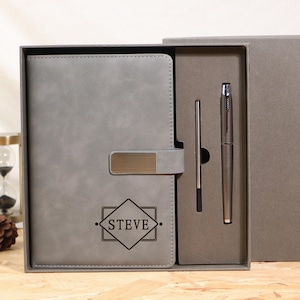 May include: A gray notebook and pen set in a gift box. The notebook has a metal clasp and the name "STEVE" inside a diamond-shaped design. The pen and stylus are silver-colored.