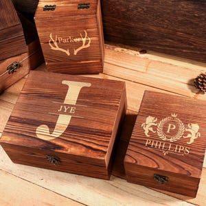Può includere: Diverse scatole di legno con incisioni personalizzate. Una scatola presenta il nome "Parker" con un design a corna. Un'altra scatola mostra la lettera "J" con il nome "JYE". Una terza scatola ha il nome "Phillips" con un stemma.