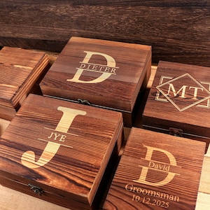 May include: Several wooden boxes with engraved initials and names. The boxes are a warm brown color with a natural wood grain. One box reads "Dieter", another "JYE", and another "MT". One box reads "David Groomsman 10.12.2025".