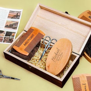 Puede incluir: Un juego de caja de madera que contiene herramientas de aseo. El juego incluye un peine, tijeras, un cepillo y un cepillo ovalado, todos de madera. El peine y el cepillo ovalado están grabados con el nombre "Michael". La caja está abierta, revelando el contenido.