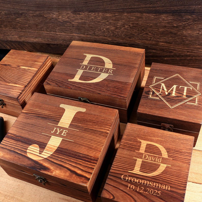 May include: Several wooden boxes with engraved initials and names. The boxes are a warm brown color with a natural wood grain. One box reads "Dieter", another "JYE", and another "MT". One box reads "David Groomsman 10.12.2025".