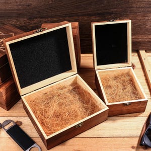 May include: Wooden gift boxes with open lids. The boxes are stained a dark brown color and have a light brown interior. One box has a black interior. The boxes are filled with a light brown packing material.