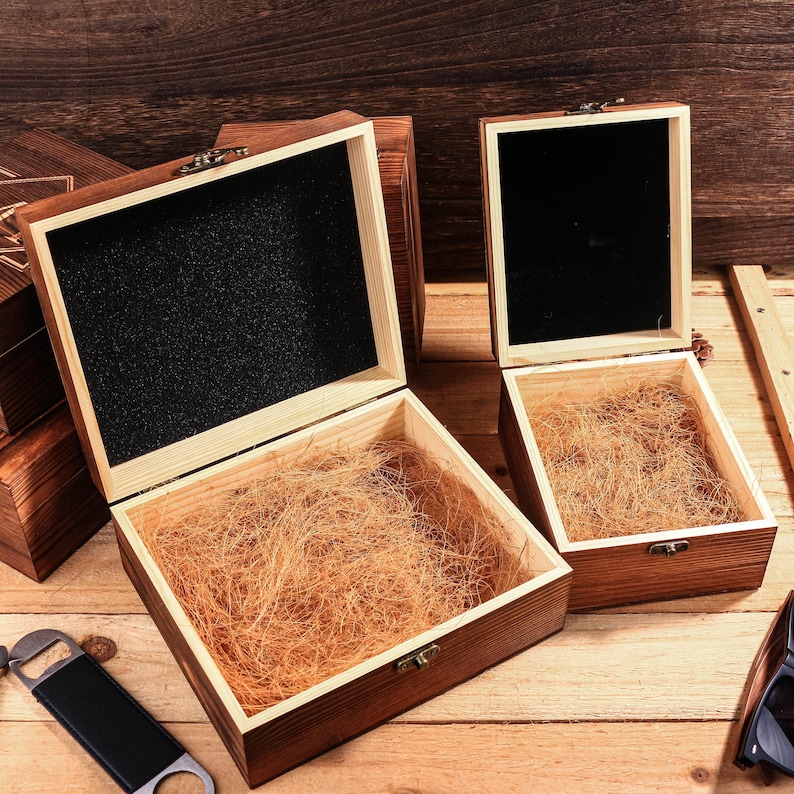 May include: Wooden gift boxes with open lids. The boxes are stained a dark brown color and have a light brown interior. One box has a black interior. The boxes are filled with a light brown packing material.