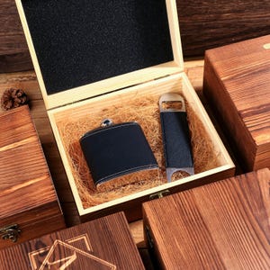 May include: A wooden gift box with a black leather flask and bottle opener. The flask has white stitching and a silver cap. The box is lined with straw and has a black foam insert in the lid. Other wooden boxes are visible.