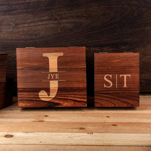 May include: Two wooden boxes with a dark brown finish. The larger box has a large letter "J" with "JYE" underneath. The smaller box has the letters "SIT". Both boxes are on a wooden surface.