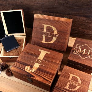 May include: Wooden gift boxes with personalized initials and names are displayed. One box is open, revealing a flask. The boxes are various sizes and have a rustic, natural wood grain finish. The initials and names are engraved.