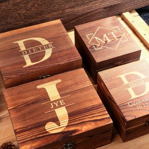 May include: Four wooden boxes with personalized engravings. The boxes are a warm brown color with a visible wood grain. Each box features a different initial and name or initials, with a decorative font. The boxes are closed with metal clasps.