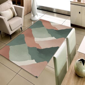 May include: A rectangular rug with an abstract design in shades of dusty rose, sage green, and off-white. The rug's pattern consists of overlapping shapes. It is placed on a tiled floor, with a chair and table visible in the background.