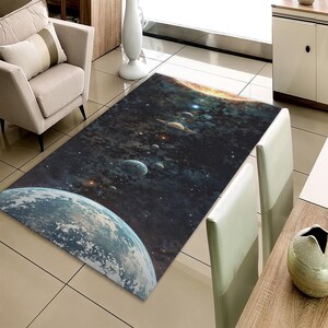 May include: A rectangular area rug featuring a detailed depiction of space, including planets, stars, and galaxies. The rug's design showcases a vibrant color palette of blues, greens, and yellows, creating a cosmic scene. The rug is placed in a living room setting.