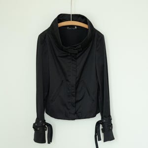 May include: A black jacket with a high, rounded collar and long sleeves. The jacket features a button closure and two front pockets. The cuffs have decorative straps with metal accents. The jacket is hanging on a wooden hanger.