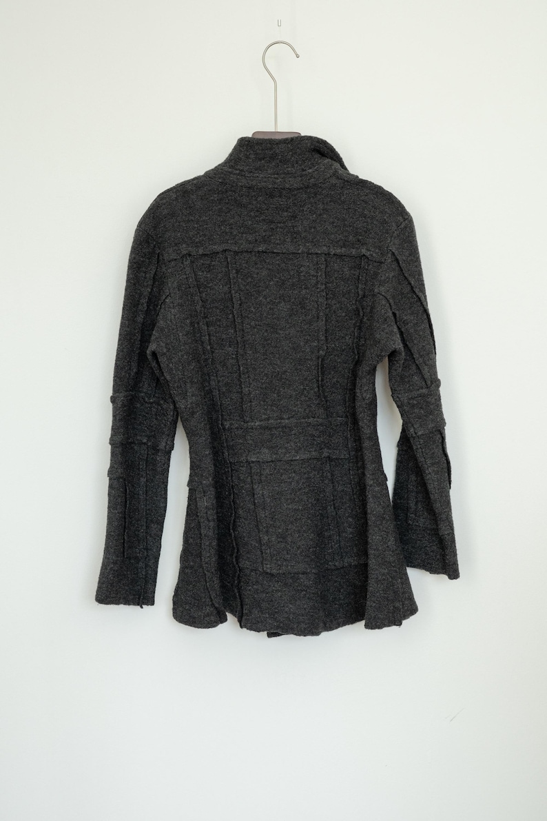 Peut inclure: Un manteau en laine gris fonc&eacute; avec un col montant et des manches longues. Le manteau a une coupe ajust&eacute;e avec des coutures et un bas &eacute;vas&eacute;. Il est suspendu &agrave; un cintre en m&eacute;tal sur fond blanc.