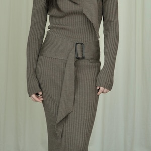 Op de afbeelding: Olijfgroene geribbelde gebreide jurk met lange mouwen en een off-the-shoulder ontwerp. De jurk heeft een bijpassende riem met een rechthoekige zwarte gesp en een gedrapeerd stoffen detail. De jurk is knielang.
