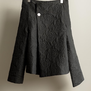 May include: A black quilted skirt with a floral pattern, hanging on a wooden hanger. The skirt has a button closure and a flared silhouette. The fabric appears to be a soft, matte material.