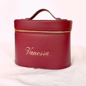 Pode incluir: Uma nécessaire bordô com zíper e alça dourados. O nome "Vanessa" é bordado em letras douradas na frente. A nécessaire tem formato oval e parece ser feita de couro texturizado.