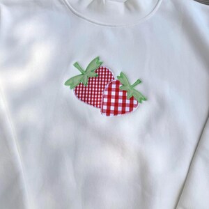 May include: A white long-sleeve sweatshirt featuring a central applique of two strawberries. The strawberries are crafted from red and white gingham fabric with light green fabric leaves stitched onto the front of the garment.