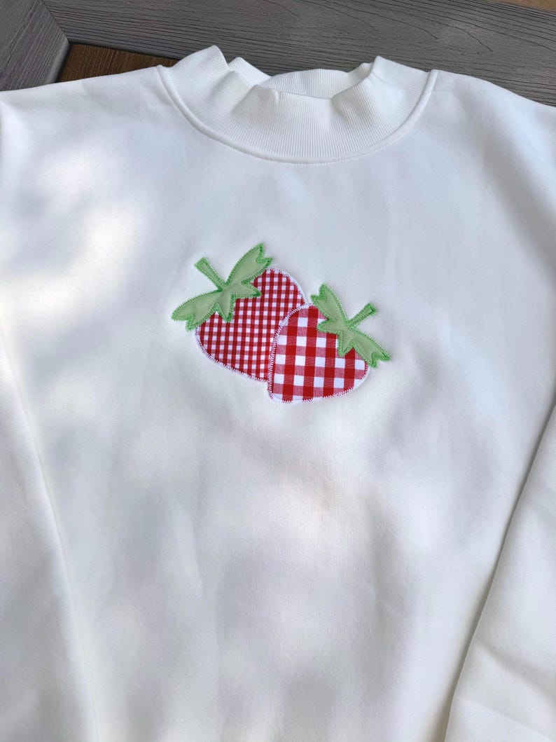 May include: A white long-sleeve sweatshirt featuring a central applique of two strawberries. The strawberries are crafted from red and white gingham fabric with light green fabric leaves stitched onto the front of the garment.