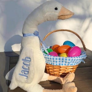 Puede incluir: Un ganso de peluche blanco con el nombre "Jacob" bordado en azul. Junto a él, una cesta de mimbre llena de coloridos huevos de Pascua de plástico. La cesta tiene un forro a cuadros azules y blancos y un asa trenzada.