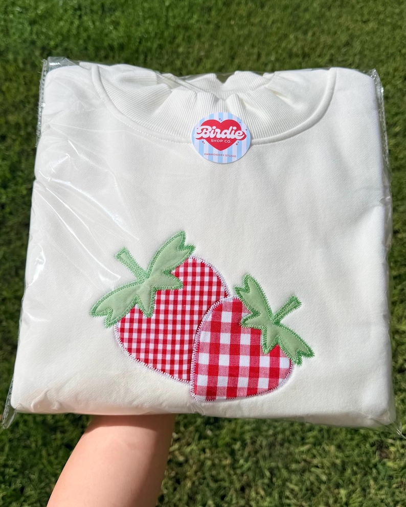 May include: A white crewneck sweatshirt featuring an embroidered applique of two strawberries with a red and white gingham pattern and light green leaves. A circular sticker with the text Birdie Shop Co. is attached to the neckline.